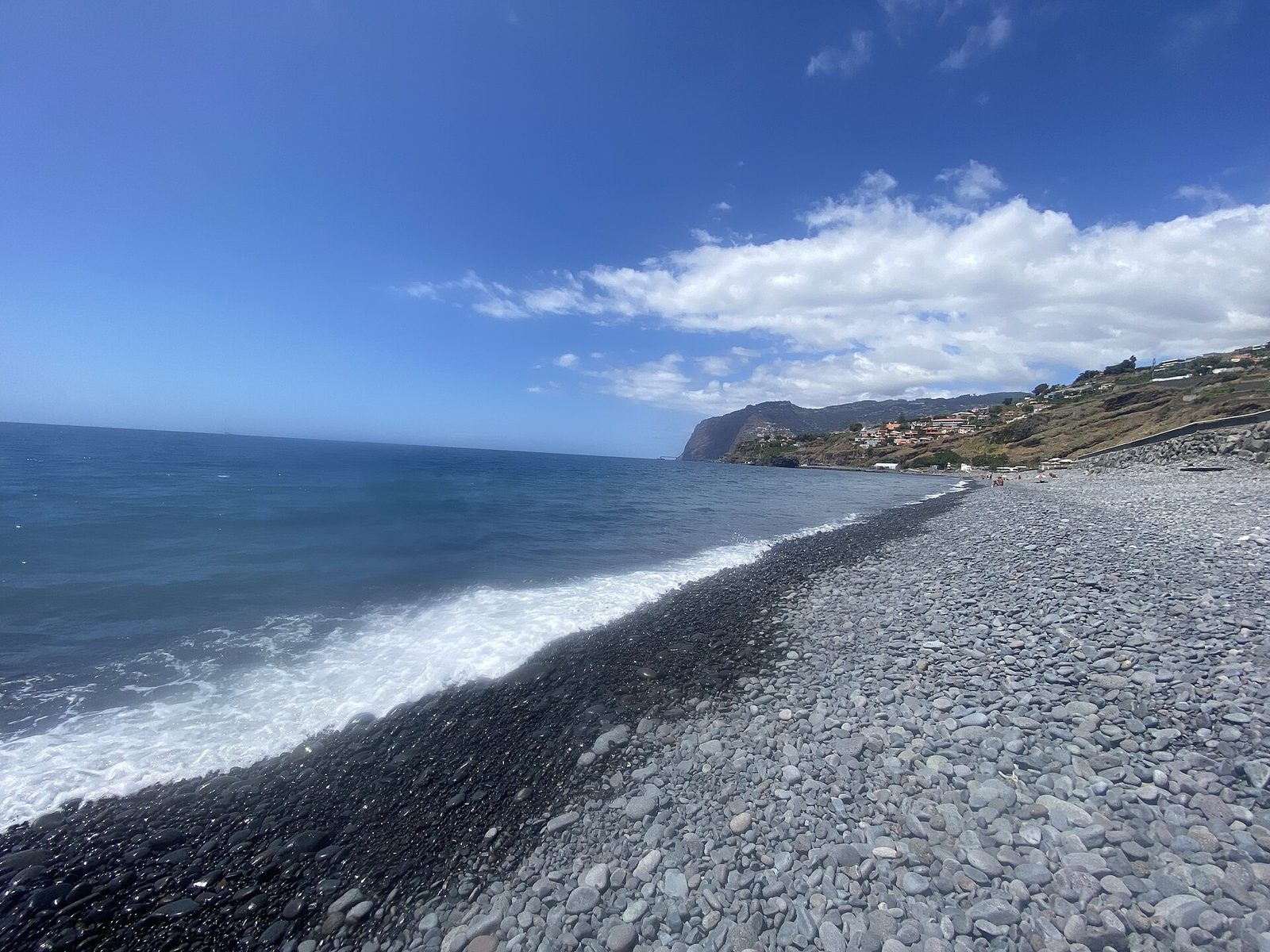 A long shore of grey rounded cobbles falling to a strip of wet black sand at the surf line, flat blue sea to the left and a distant headland with hillside houses.