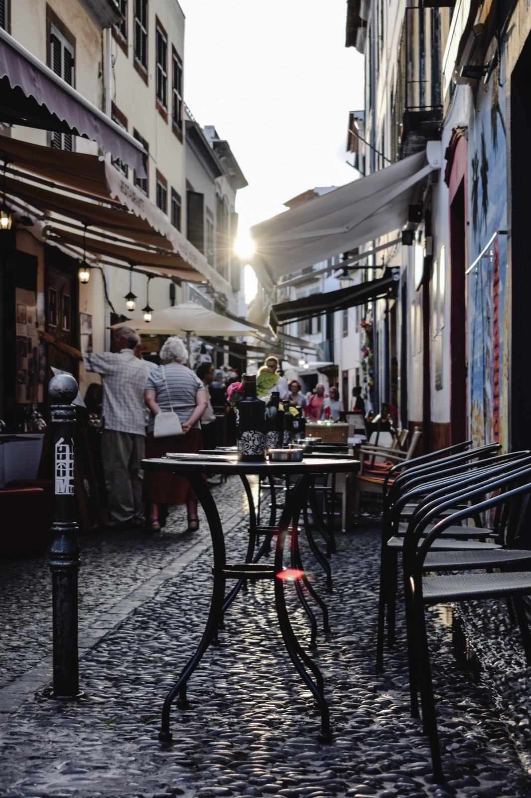 A narrow cobbled lane in Funchal’s Old Town at low sun, restaurant tables set out on the paving, awnings overhead and painted panels on the wall.