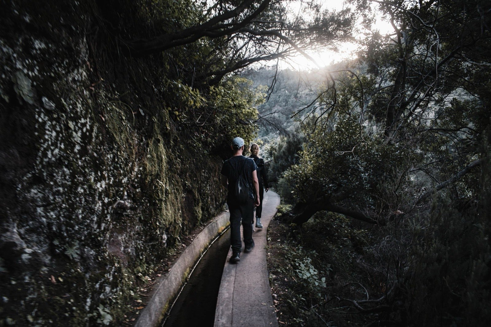 Two walkers following a levada path into low sun, the narrow water channel running full alongside the trail, a mossy rock face on one side and a wooded valley dropping away on the other.