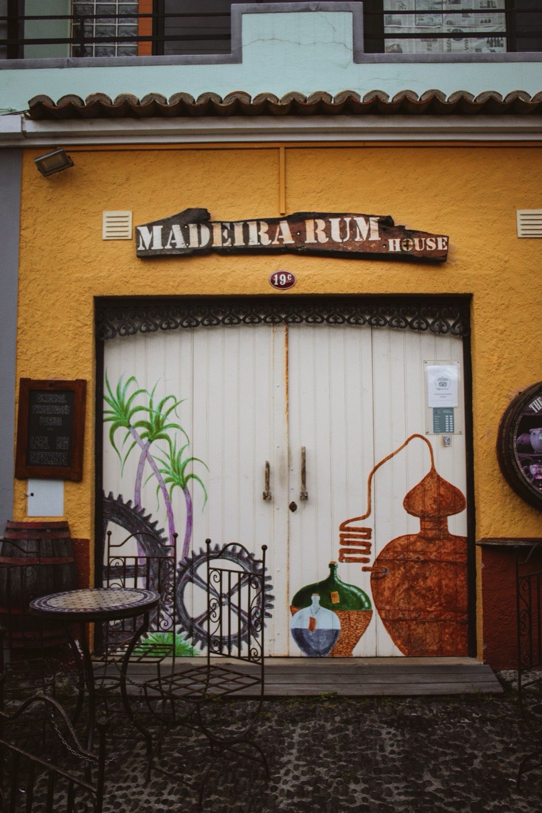 The closed white double doors of a Funchal rum bar on a mustard-yellow wall, painted with sugar cane, a copper still and a demijohn, a cafe table on the cobbles outside.
