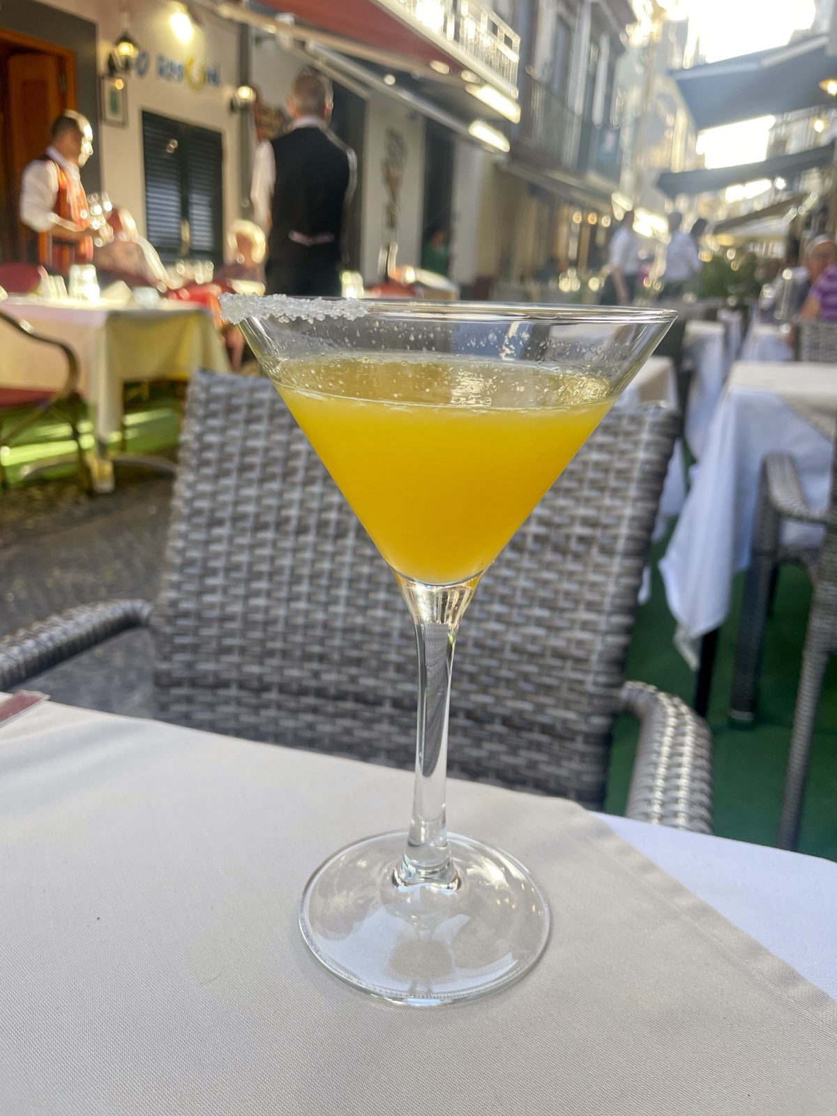 A sugar-rimmed stemmed glass of orange poncha on a white tablecloth at a pavement restaurant in Funchal, with waiters and lit awnings behind.