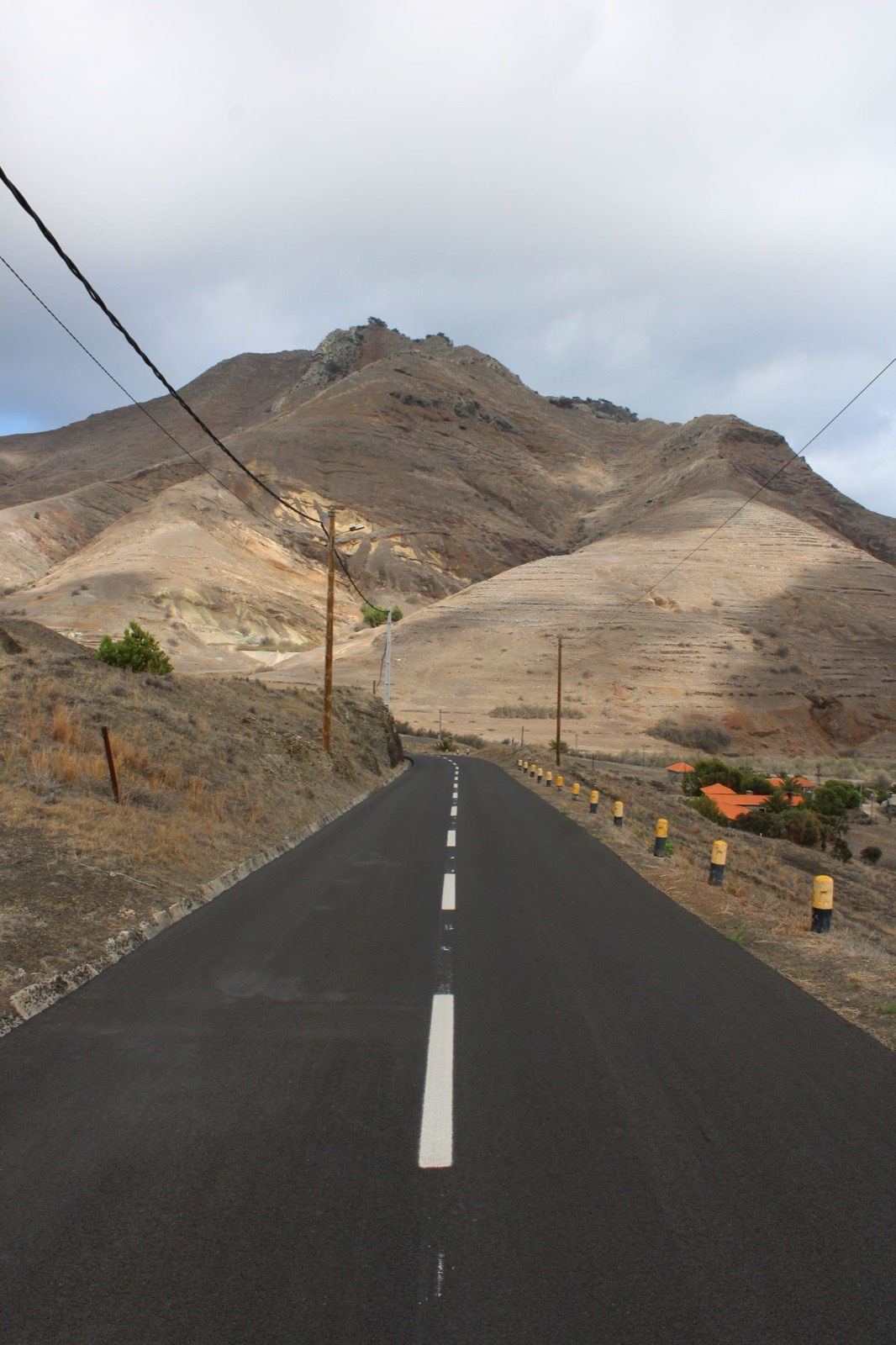 An empty two-lane road running dead straight towards bare, dust-coloured hills on Porto Santo, with overhead cables, a few pines and orange-tiled roofs at the foot of the slope.